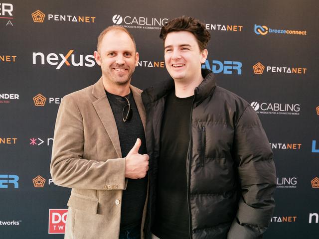 Two men in front of a Pentafest sponsors board, one is giving a thumbs up